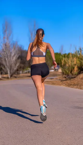 Attractive young runner girl, dressed in tight running shorts and top, running on an asphalt path through a mountain park illuminated by dawn sunlight.
