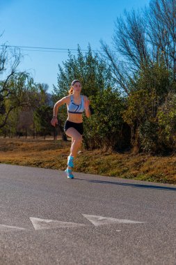 Koşu şortu ve beyaz bir bluzla koşan mutlu genç bir koşucu kız, stil ve mükemmel teknik hareketlerle koşuyor bir doğa parkında sabah ışığında asfalt bir yolda..