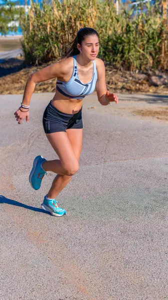 A young female runner, dressed in short running pants and a top, positioned with style and perfect technical movement with her legs bent and body tilted ready to sprint on an asphalt path in a park.