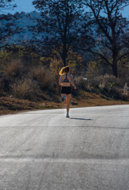 Şafak sökerken arka planda kalan genç bir koşucu kız bir doğa parkında asfalt üzerine gölgesini düşürerek havada asfalt bir şekilde asılı duruyor..