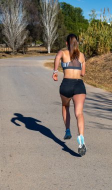 Koşu şortu ve dar spor bluzu giyen genç koşucu kız, asfalt yolda bir adım atarak havada asılı duruyor ve uzun gölgesini yere yansıtıyor..