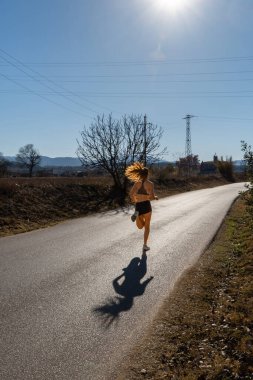 Dar bir şort ve dar bir tişört giymiş, bir doğa parkında asfalt bir yolda koşan, güneş ışınlarıyla gölgesini düşüren zinde genç bir koşucu kadın..