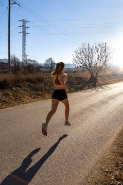 Dar bir şort ve dar bir spor bluz giymiş zinde genç bir bayan koşucu, arka plandaki şafak ışığına doğru zarif bir şekilde koşuyor. Bir doğa parkında asfalt bir yolda gölgesini bırakıyor..