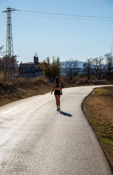 Şafak sökerken arka planda genç bir koşucu kız asfaltta gölgesini bırakarak doğal bir parkta asfalt bir yolda rahatça yürüyor..