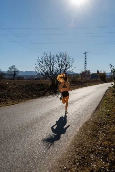 Dar bir şort ve dar bir tişört giymiş, bir doğa parkında asfalt bir yolda koşan, güneş ışınlarıyla gölgesini düşüren zinde genç bir koşucu kadın..