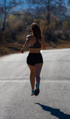 Genç kaslı kadın koşucu, dar şortlu ve üstsüz, asfalt bir yolda havada asılı, güneş ışığıyla aydınlanan bir dağ parkında gölgesini düşürerek koşuyor..