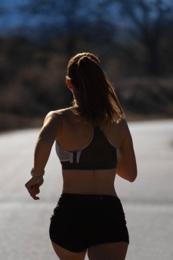 Arkadan, dar şortlar ve üstten giyinmiş, asfalt bir yolda koşan genç bir kadın koşucunun arka ışıklandırılmış silueti bir doğa parkında gün ışığıyla aydınlatılıyor..