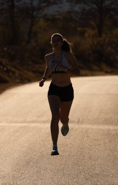 Kaslı koşucu kız silueti, dar şortlu ve üstsüz, asfalt bir yolda asfalt yolda asılı, şafak vakti aydınlanan bir dağ parkından geçiyor..