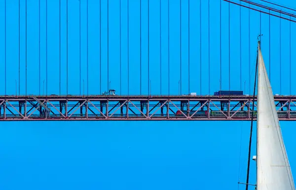 Top of the white sail of a sailboat sailing in front of the red steel 25 de Abril suspension bridge with road traffic and a train traveling inside the bridge under a clear blue sky.