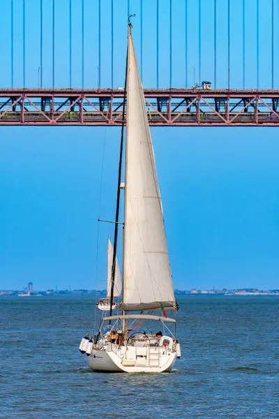 Two tourist recreation sailboats sailing on the Tagus River with the red steel 25 de Abril suspension bridge above them with car and truck traffic and a train circulating inside the bridge.