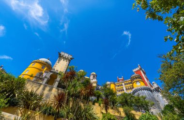 Pena Sarayı 'nın geniş açılı dış manzarası mozaikler ve güçlendirilmiş duvarlar ve güneşli mavi gökyüzünün altındaki palmiye ağaçları ile süslenmiş sarı ve kırmızı boyalı duvarlar. Sintra. Portekiz.