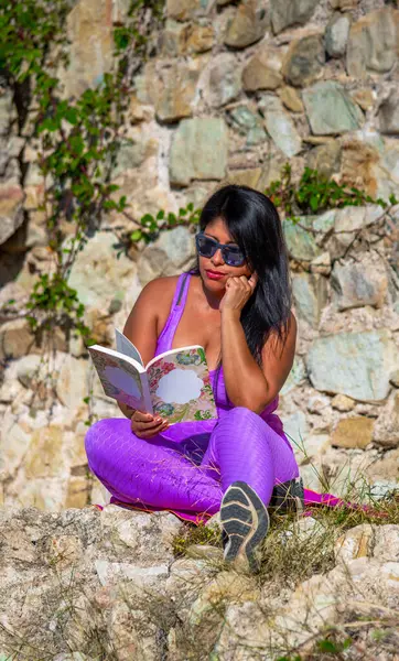Sexy dark-skinned Latin reading woman, dressed in pink tight leggings and top, reading concentratedly, in sunglasses, a self-help book, sitting in the field in the ruins of a castle.