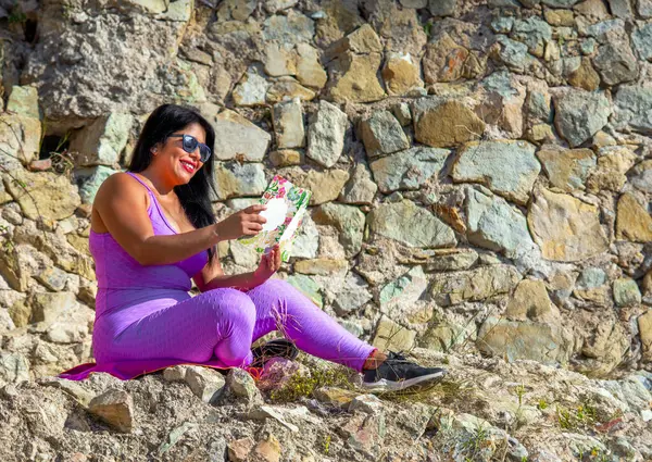 Sexy Latin woman with dark skin, dressed in pink tight leggings and top, happily and cheerfully reading a self-help book, sitting in the field in the ruins of a castle with rural background.