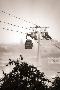 Bulutlu bir gökyüzünün altından sarkan çelik kabloların üzerinde asılı duran bir Vilanova de Gaia teleferiği. Klasik Gaia mahallesi sisle örtülü. Siyah ve beyaz.