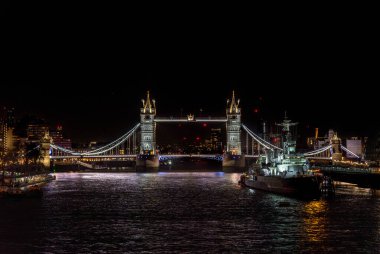 Tower Bridge 'in önü gece aydınlandı ve savaş müzesi gemisi HMS Belfast Thames Nehri' ne yanaştı. Sudaki restoranların ve binaların ışıklarının yansımalarıyla..