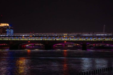 Gece vakti Londra 'daki Blackfriars Köprüsü' ne park eden gece treninin arka planında Thames Nehri ve Milenyum Köprüsü 'nün suyuna yansıyan menekşe ışıklarıyla yayalar yürüyordu..