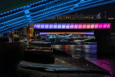 Perçinlerin ayrıntıları ve Thames nehrinin kıyılarını aydınlatan bir Londra köprüsünün çelik yapısı. Fütürist mavi ve mor ışıklar ve diğer tarafta çalışan bir gece treni..