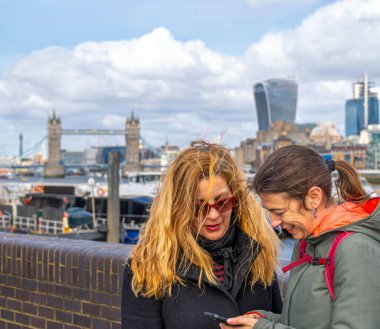 Lezbiyen bir çift mutlu bir şekilde gülümsüyor. Cep telefonlarıyla Londra 'nın merkezini arıyorlar. Arka planda Tower Köprüsü olan Thames nehrinden..