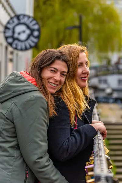 Stock image A lesbian couple hugging and happy leaning on the fence of Camden Lock Regent Canal Bridge with love padlocks closed on the fence in the Camden Town neighborhood of London.