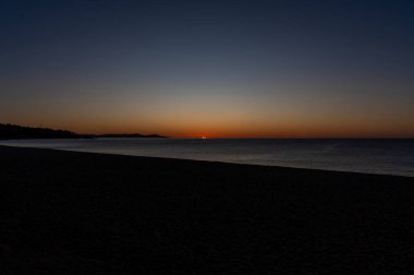First rays of the sun emerging on the sea horizon line at dawn illuminating with sunlight mirage in the water of the coast of the calm Mediterranean Sea, with the sky tinted orange and blue. clipart