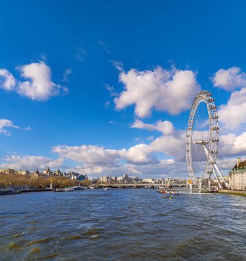 Thames nehrinin panoramik manzarası gün batımında ışıldıyor ve London Eye, dünyanın en büyük ve en uzun dönme dolabı, arka planda, mavi bulutların altında.