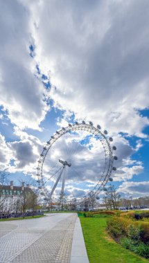 Yeşil çimenli bir parkın arka planında büyük bir İngiliz dönme dolabı beyaz bulutlarla mavi bir gökyüzünün altında yürüyor. Londra, İngiltere, Birleşik Krallık.