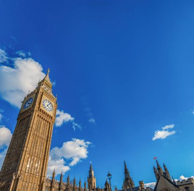 Büyük Ben Saat Kulesi ve Westminster Manastırı 'nın alçak açılı görüntüsü açık mavi gökyüzü ve beyaz bulutlarla aydınlık bir günde akşam güneşiyle aydınlanır. Londra.
