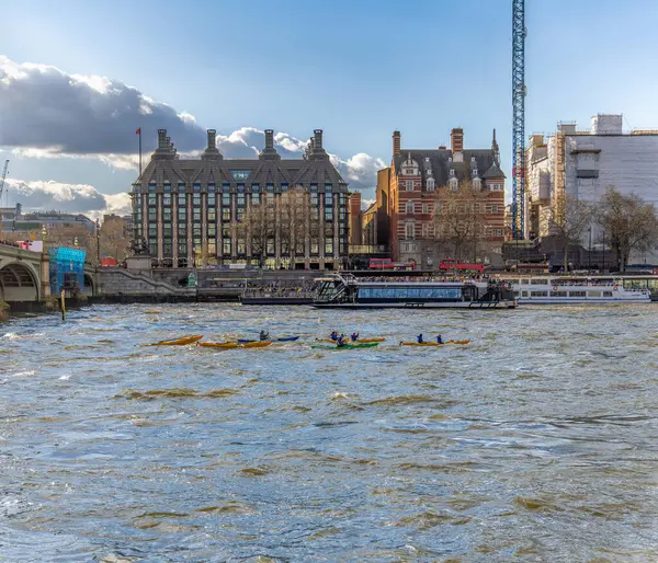 Arkadaşları ve ailesi Thames nehrinde kano ve kayıklarla su sporu yapıyor. Arka planda feribot ve turistik tekneler var..