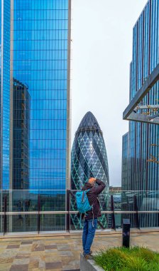 Orta yaşlı bir fotoğrafçı bir gökdelenin çatısından neo-fütürist gökdelene fotoğraf çekiyor. 30 St. Mary Axe 'de kaldırma platformunda cam cephe temizleyen bir işçi var..