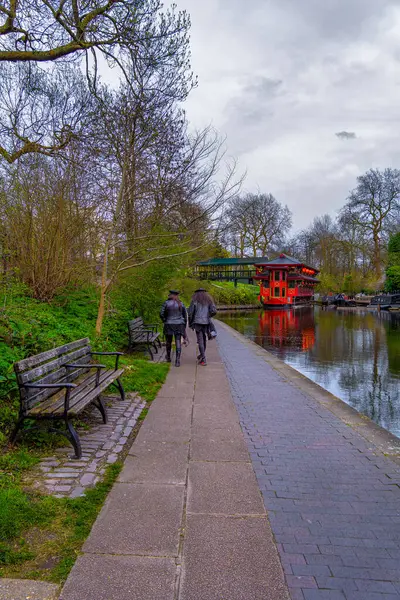 stock image Two friends in dark gothic leather clothing strolling on the Regent Canal promenade with a red wooden floating Chinese restaurant moored to it.