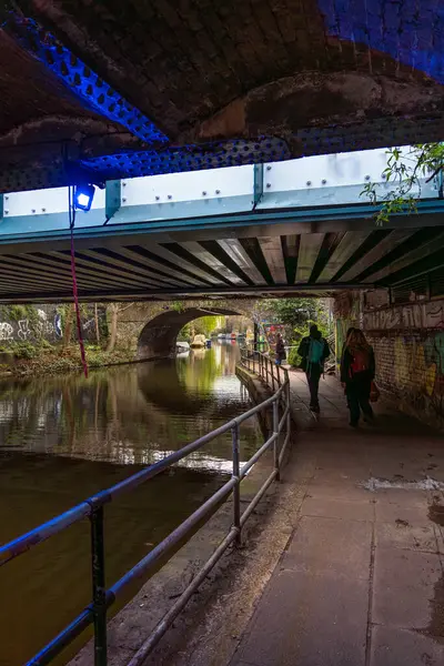 stock image Friends walking sightseeing under a Regent's Canal promenade bridge with moored houseboats and graffiti on the walls.