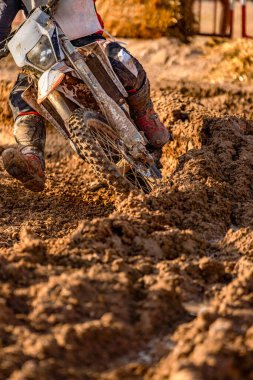 Bir off-road motosikletinin ön kısmı virajda sürerken çamurlu arazide tekerleklerle ve çamurla lekelenmiş ve kirlenmiş bir şekilde batıyor..