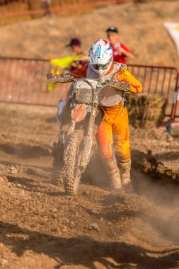 Motokros sürücüsü, arka planda iki çocuğun tezahüratıyla zorlu bir toprak yolda motorunu itiyor..