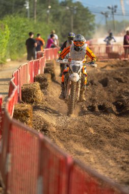 Bir off-road yarışında motokros sürücüsü ve onu yakından takip eden başka bir sürücü ve yarışı izleyen insanlar..