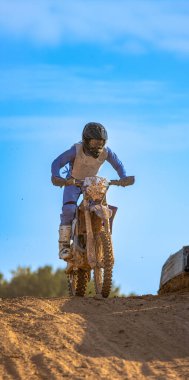 Motokros binicisi, bir tepenin üstündeki açık mavi gökyüzü altında dayanıklılık yarışında arazi motosikletinin üzerine atlamayı bitiriyor..