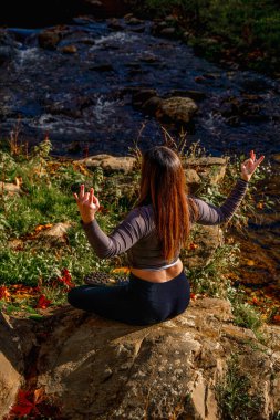 Uzun turuncu saçlı genç bir kadın Lotus pozisyonunda meditasyon yapıyor. Doğal bir ormanın kenarındaki bir kayanın üzerinde oturuyor. Gün doğumunun ışığıyla güzelce aydınlatılıyor..