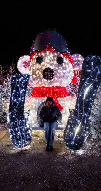 Happy woman looking at camera sitting on bench in front of giant colorful LED decoration shaped like a bear on skis on a cold night with some snow on the fir trees. clipart