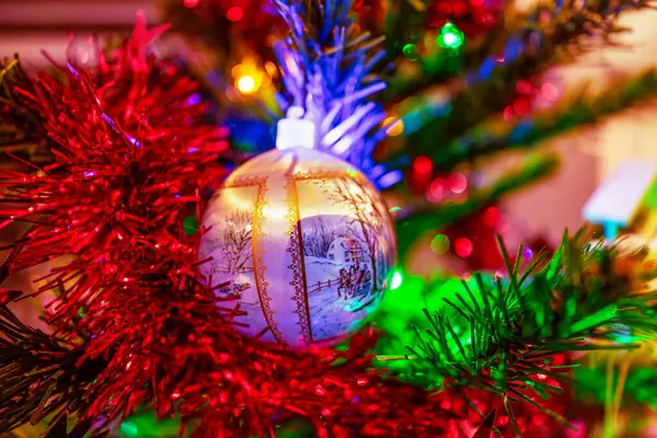 Renkli ışıklar ve çelenklerle süslenmiş ağaçta asılı duran parlak bir Noel süsü içinde karlı bir yolda giden bir araba tasviri.