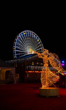 LED ışıklardan yapılmış aydınlanmış Noel meleği trompet çalıyordu. Gece bir şenlik gece pazarında aydınlanmış bir dönme dolabın köşkünden çıkmış gibi görünüyordu..