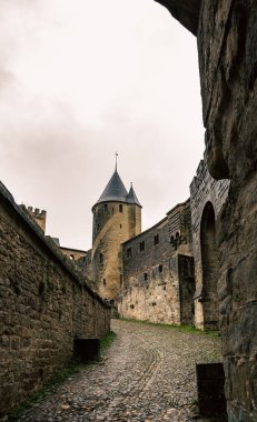 Uphill cobbled street in the City de Carcassonne, France, with a view of a medieval castle tower on a cloudy day with damp ground. clipart