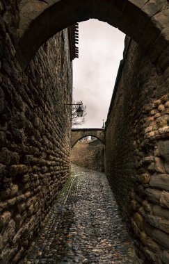 Fransa 'nın Carcassonne şehrinde bulutlu bir günde, üzerinde taş kemerler ve dövülmüş demir fenerler asılı duran kaldırım taşından bir sokak..