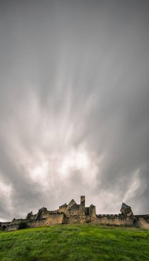 Medieval fortress of Carcassonne with stone walls and towers seen from a low angle from a hillside with wet green grass under a spectacular cloudy sky. clipart