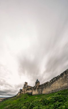 Vertical panoramic view of the historic medieval Carcassonne Castle with stone walls and towers from a hillside with wet green grass under a cloudy grey sky on a rainy day. clipart