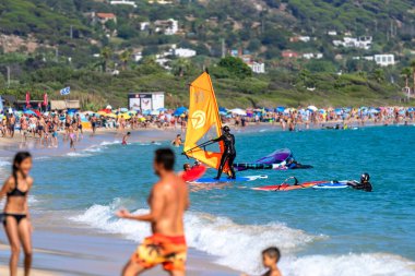 Rüzgâr sörfçüleri, arka planda tatil evleriyle birlikte Tarifa, İspanya 'da güneşli bir günde oynayan ve eğlenen hamamlar ve ailelerle dolu bir plaja geliyorlar..
