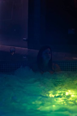 Isıtmalı bir spa havuzunda dinlenen mayo giymiş genç bir kadın ortam aydınlatması ve yanında bir bardak beyaz şarap ile..