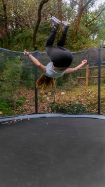 Uzun saçlı genç bir kadın ormanda trambolinde ters takla atar..