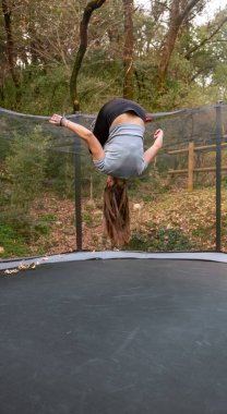 Genç, sportif bir kadın, uzun saçlarıyla bir orman parkında trambolinde yere doğru takla atıyor..