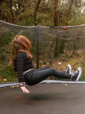 A young woman jumping sitting with her butt on an outdoor trampoline in the forest of a natural park.