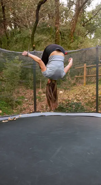 Genç, sportif bir kadın, uzun saçlarıyla bir orman parkında trambolinde yere doğru takla atıyor..