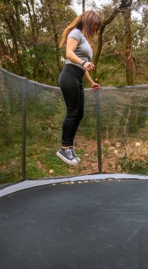 Spor kıyafetleri giymiş genç bir kadın dağlardaki açık bir ormanda trambolinde yükseğe zıplayarak anın tadını çıkarıyor..